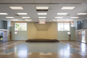 Large bright open gymnasium type room with small carpeted stage, high windows, and American flags